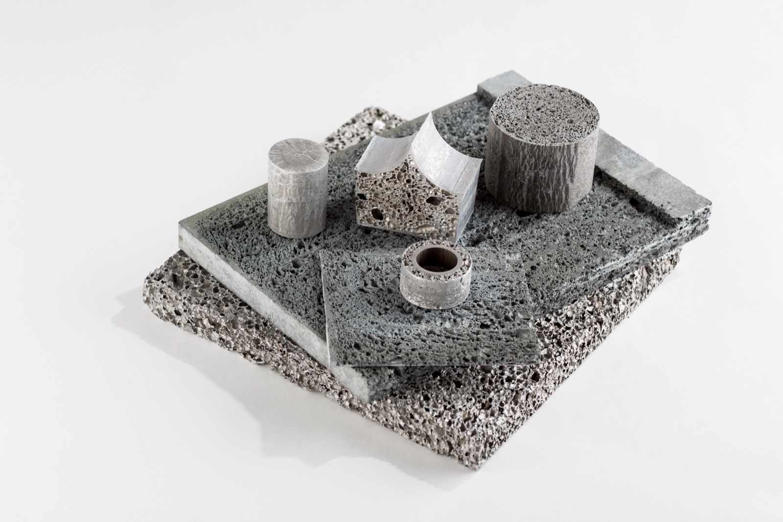 Several machined parts of aluminum foam on a foam plate: two cylinders, a ring, a piece cut into a saddle shape and a second, thinner plate. The cut faces show the cell structure throughout.
