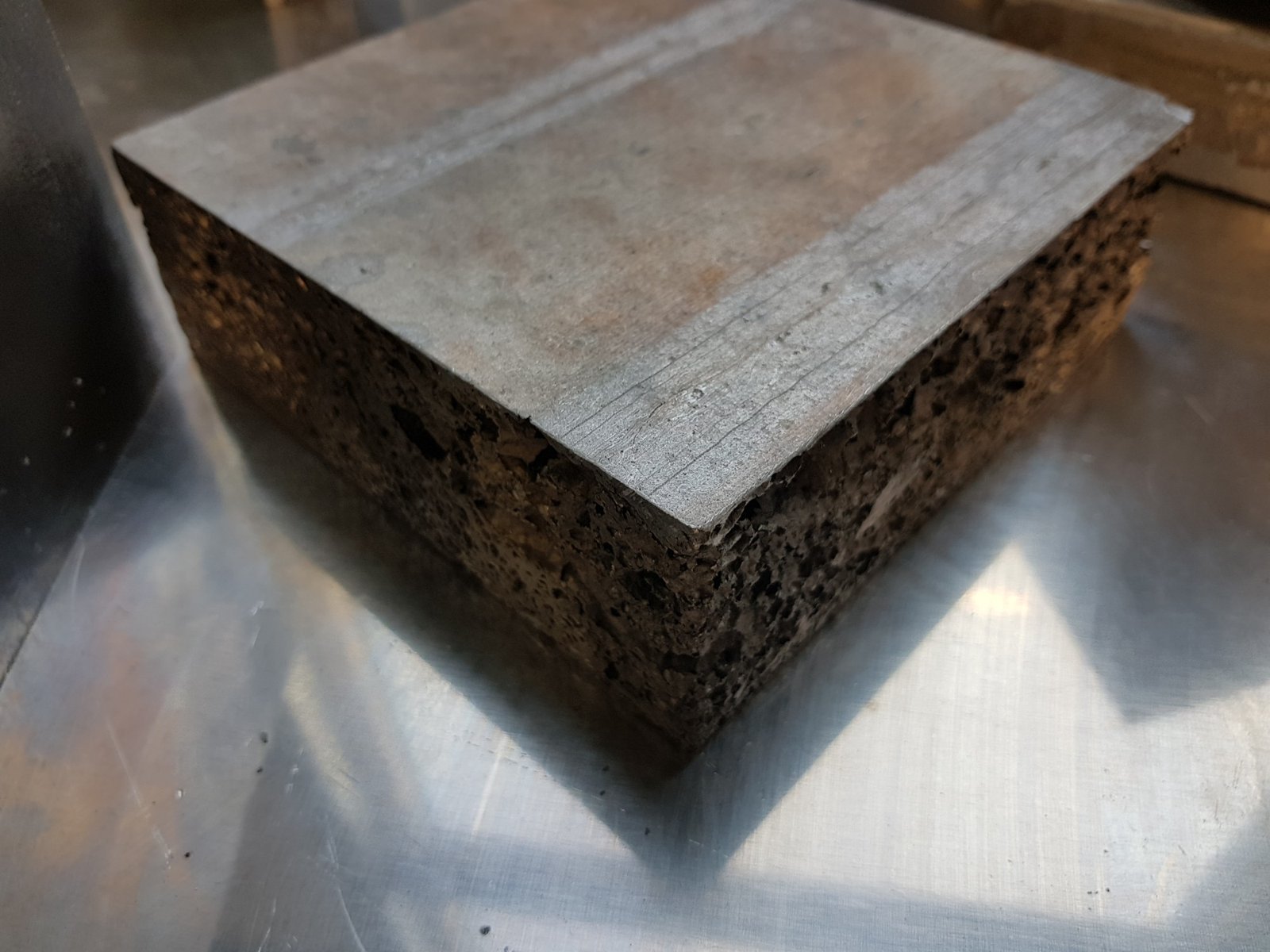A block of aluminum foam on a metal sheet: the top is a closed, smooth casting skin, and the pores show only on the machined sides.