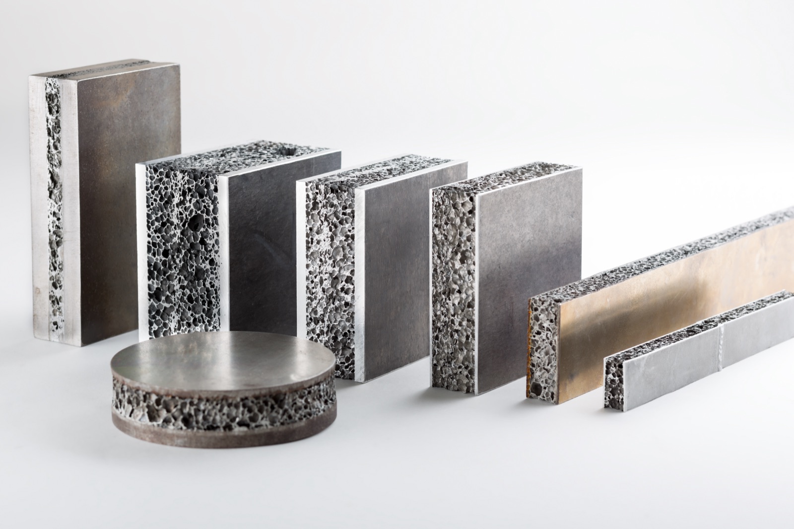Several cut sandwich blanks side by side: an aluminum foam core between light and dark cover layers, in different thicknesses, together with a round disc and two narrow strips.