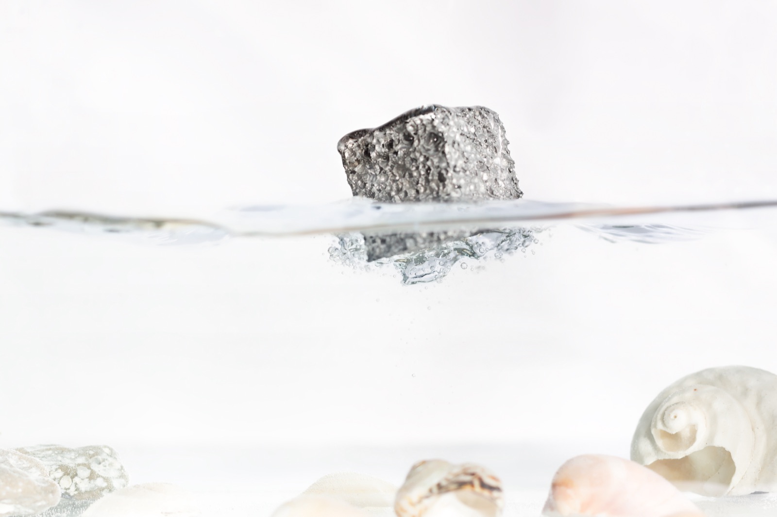 A piece of aluminum foam lies still at the water surface, most of it standing clear of the water; single air bubbles rise beneath it, and shells lie on the bottom of the basin.