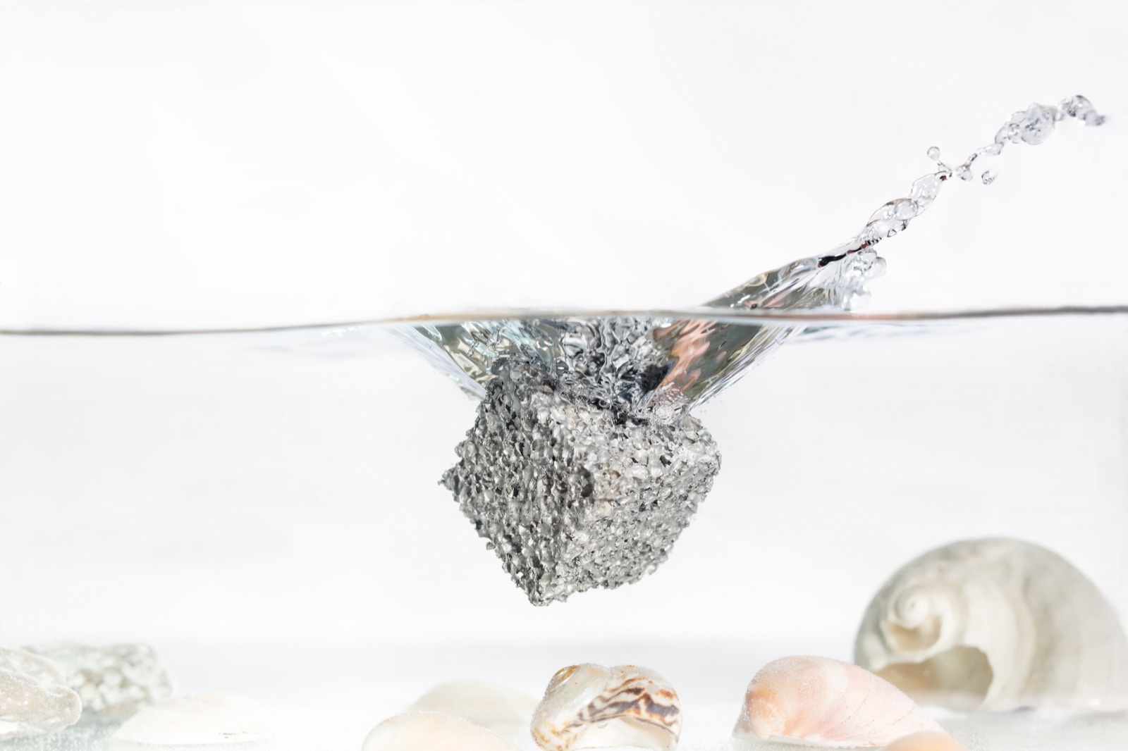 A cube of aluminum foam at the water surface, half submerged, with water splashing up; shells lie on the bottom of the basin.