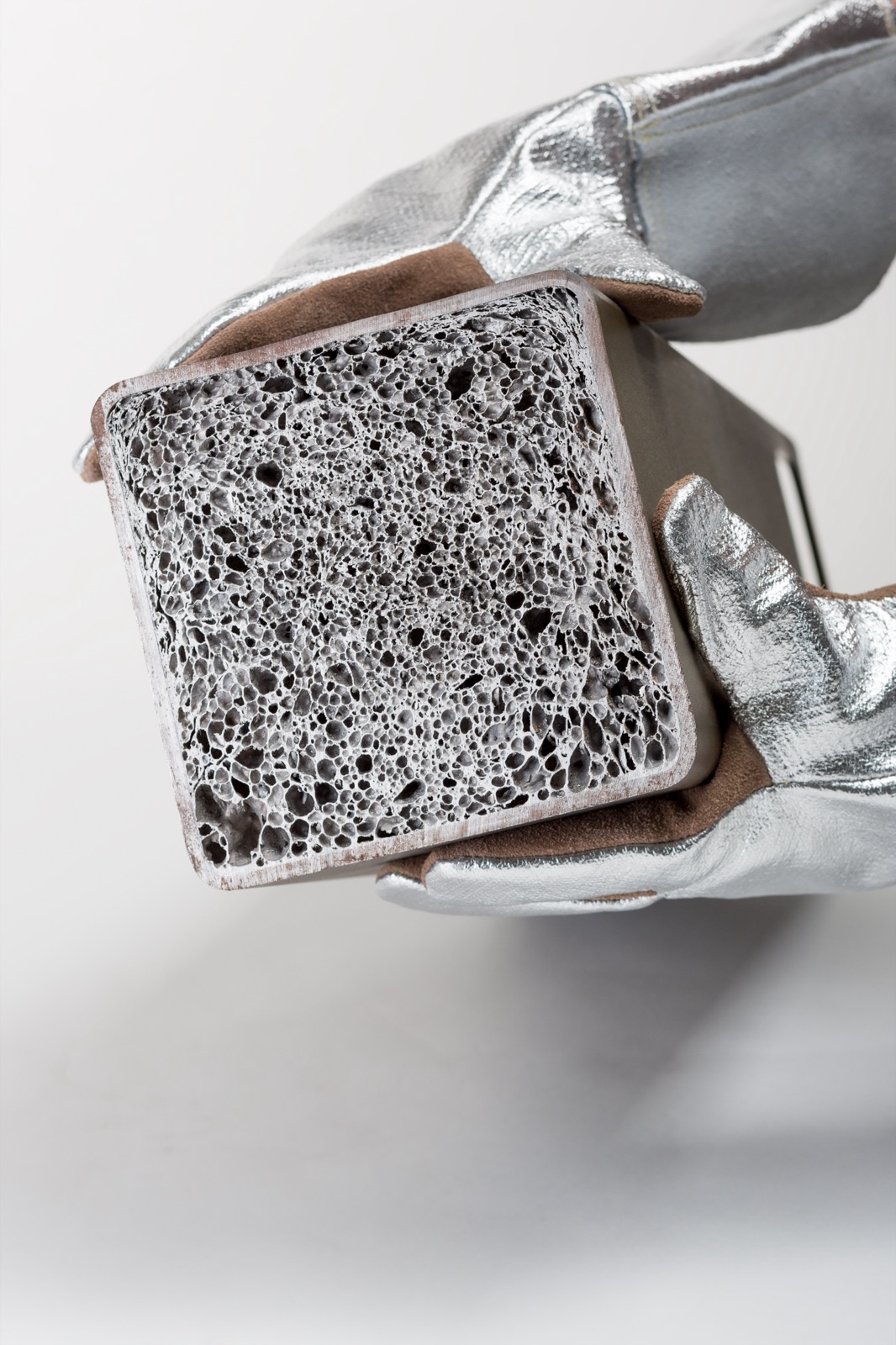 A gloved hand holds a square hollow profile with its cut face towards the viewer; the cavity is completely filled with aluminum foam, which reaches right up to the profile walls.