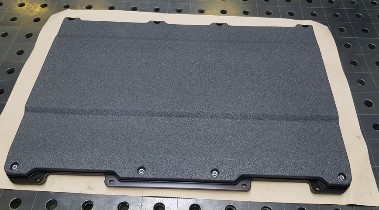 Dark grey coated protection plate with a rough surface and a bolted-on retaining strip along the lower edge, lying on a perforated table.