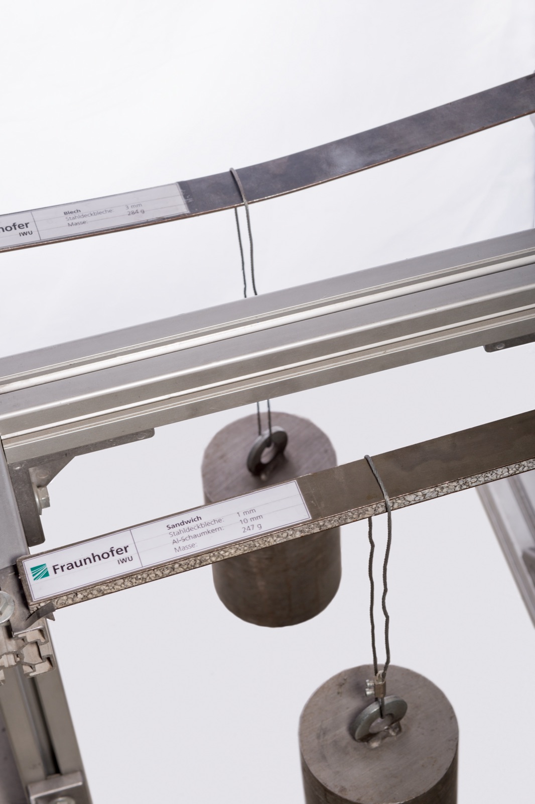 A Fraunhofer IWU demonstration set-up with two specimens clamped horizontally, each with a weight hung from it: at the top a steel sheet that bends visibly, below it a sandwich with steel cover layers and an aluminum foam core that stays straight. Labeled tags hang from both specimens.
