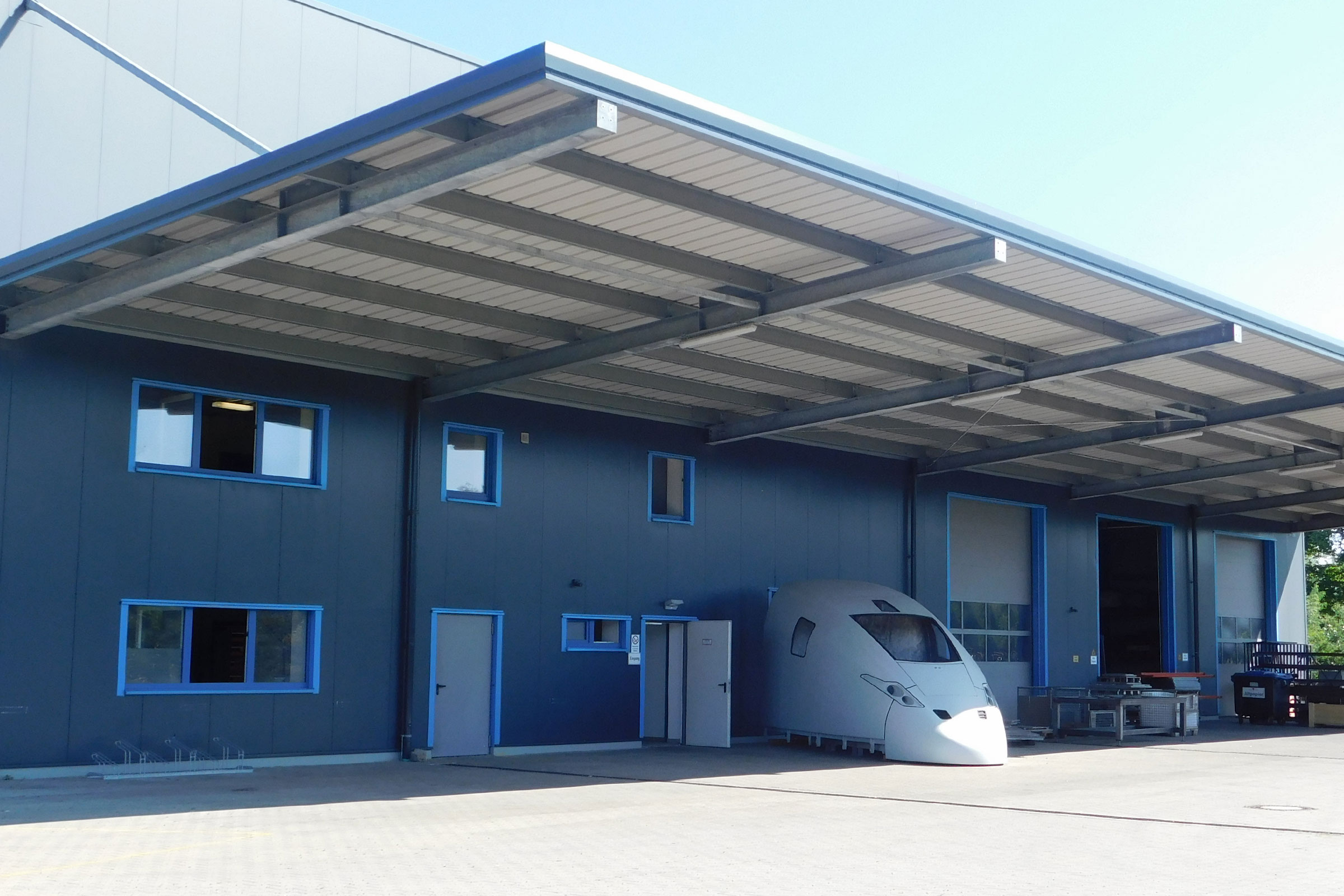 The company building with a dark blue façade, light blue window frames and a wide, projecting canopy over the hall doors; in front of it stands the unpainted power car hood of a high-speed train.