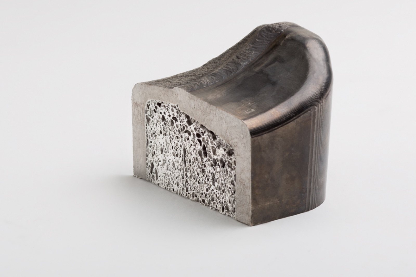 A shaped element with a saddle-shaped curved top and a cut end face where the aluminum foam core shows between the cover layers; the curved surface shows annealing tints.