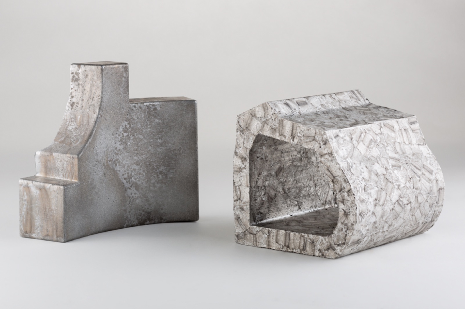 Two shaped elements of aluminum foam side by side: on the left a stepped part with a closed, smooth cast skin, on the right a part with a rectangular opening and a coarsely textured surface.
