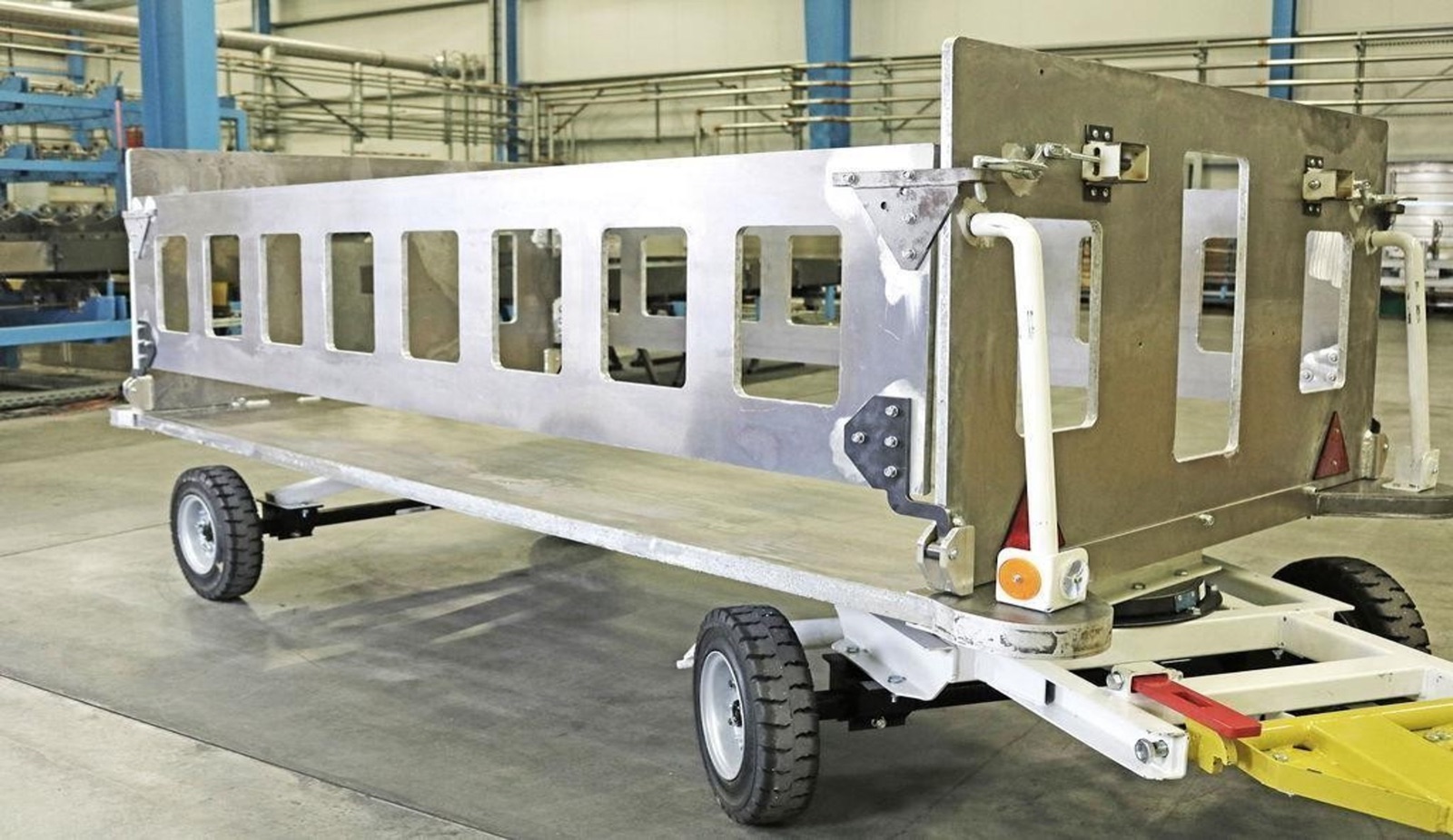 A baggage cart in a production hall: its body and side walls are made of light-colored aluminum sandwich panels with cut-out windows, mounted on a chassis with four wheels and a drawbar.