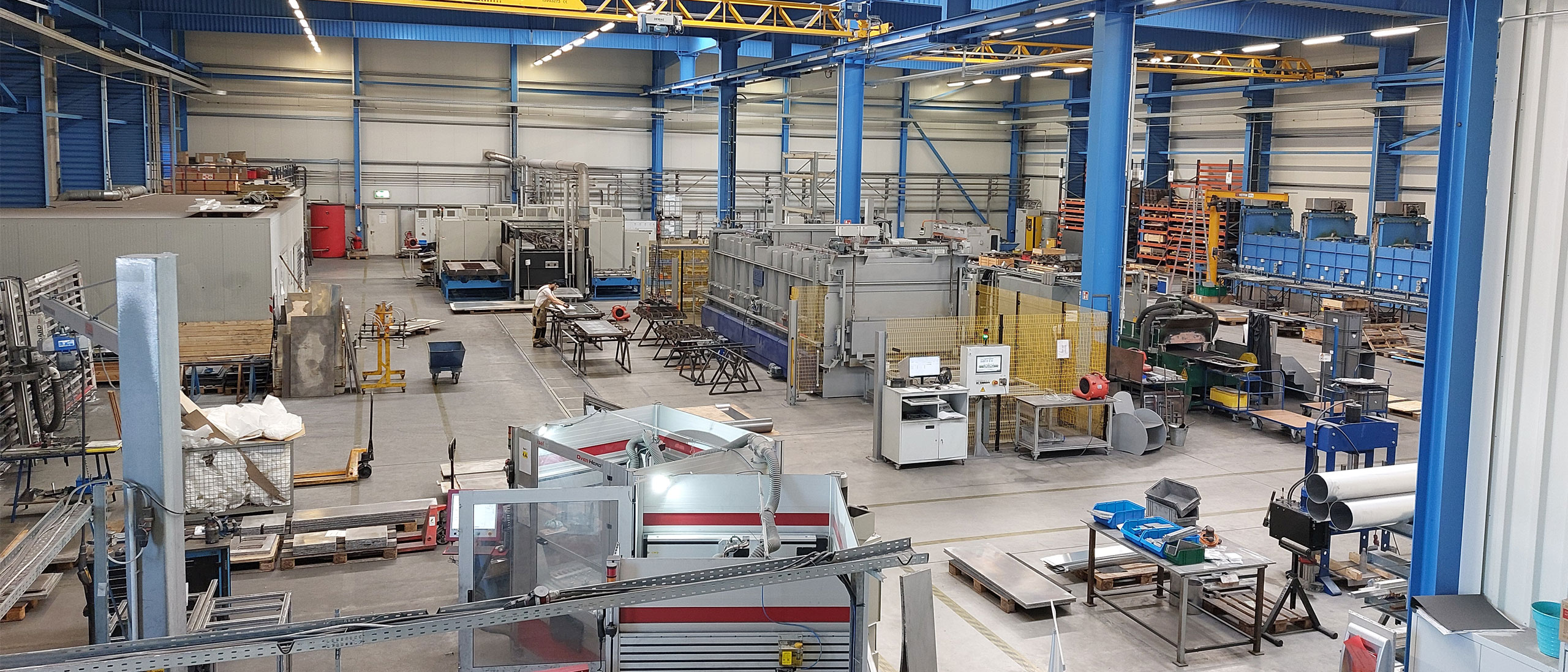 View from a gallery into a production hall with a blue steel structure and yellow overhead cranes: in the middle a long gray line behind a yellow safety fence, on the left an employee at a workbench, in the foreground machines and pallets.