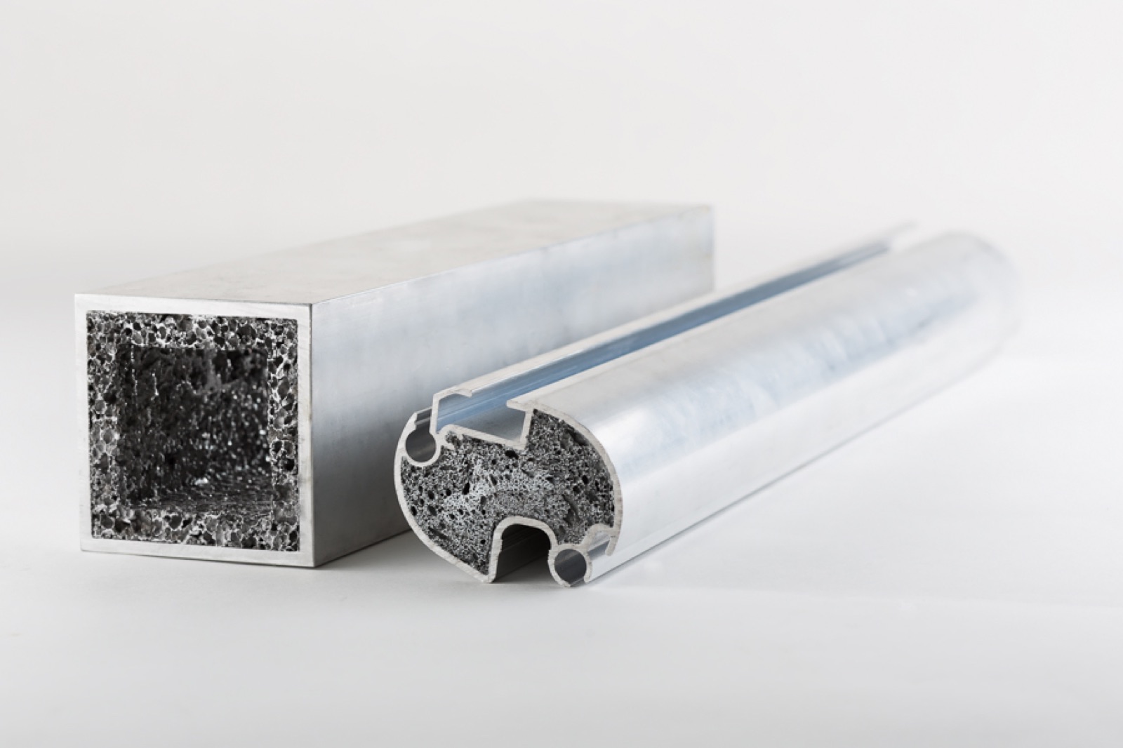 Two foamed aluminum profiles: on the left a square tube whose cut face shows a continuous layer of foam along the inner walls, on the right a grooved round profile with the foam sitting in its chamber.