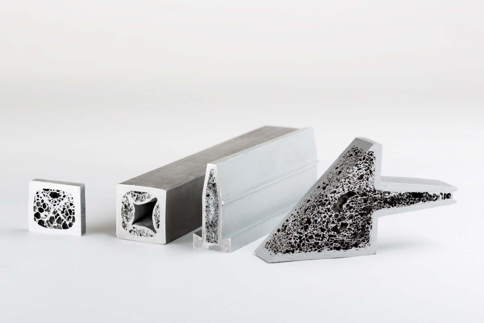 Four foamed profile sections side by side: a flat rectangle, a square tube with foamed corners, a narrow T-profile and a multi-chamber extruded profile. Every cut face shows the foam in the cavities.