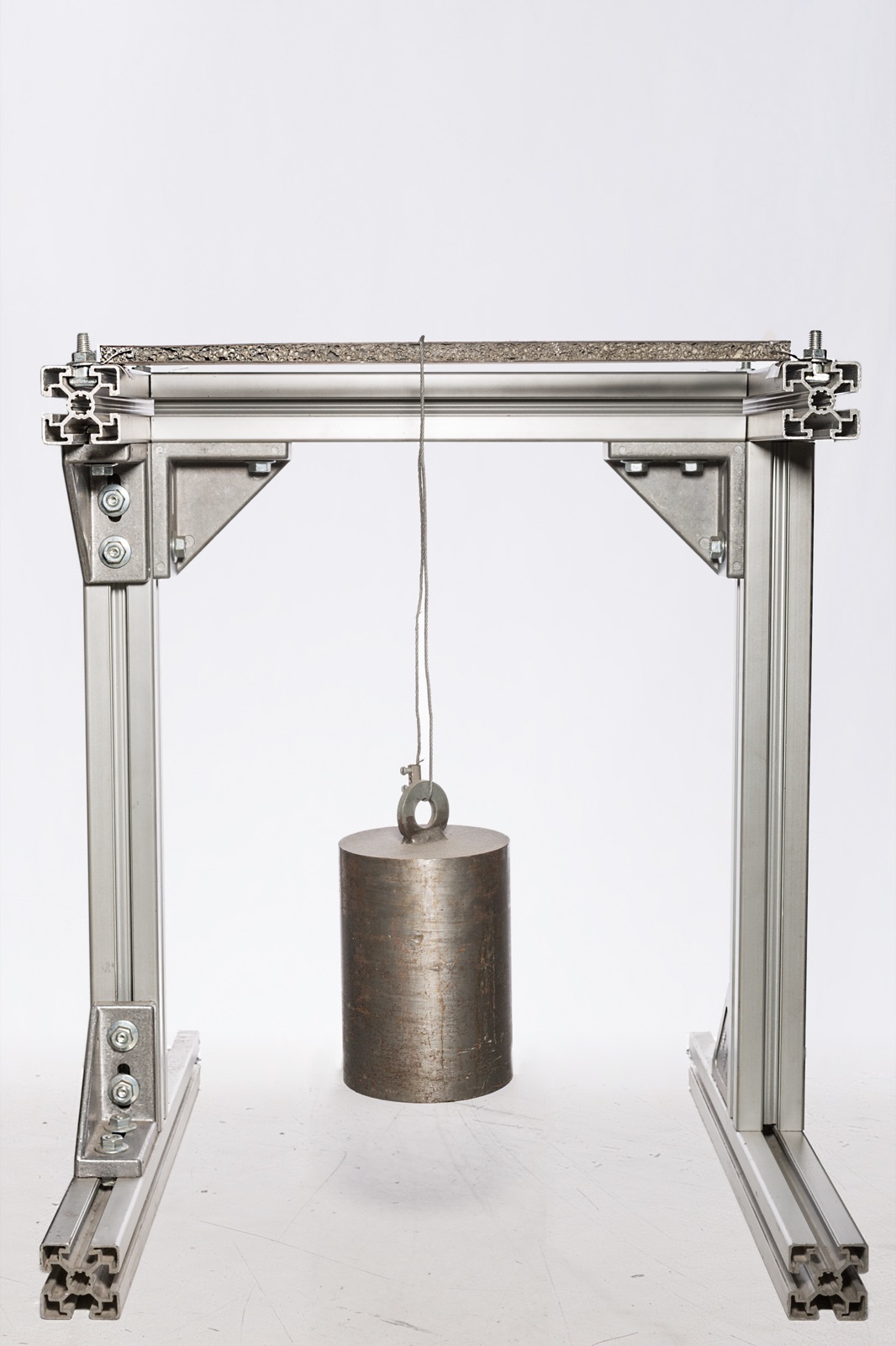 A test rig made of aluminum profiles: a sandwich specimen lies horizontally at the top, with a cylindrical steel weight hanging from its middle on a wire rope. The specimen stays straight.