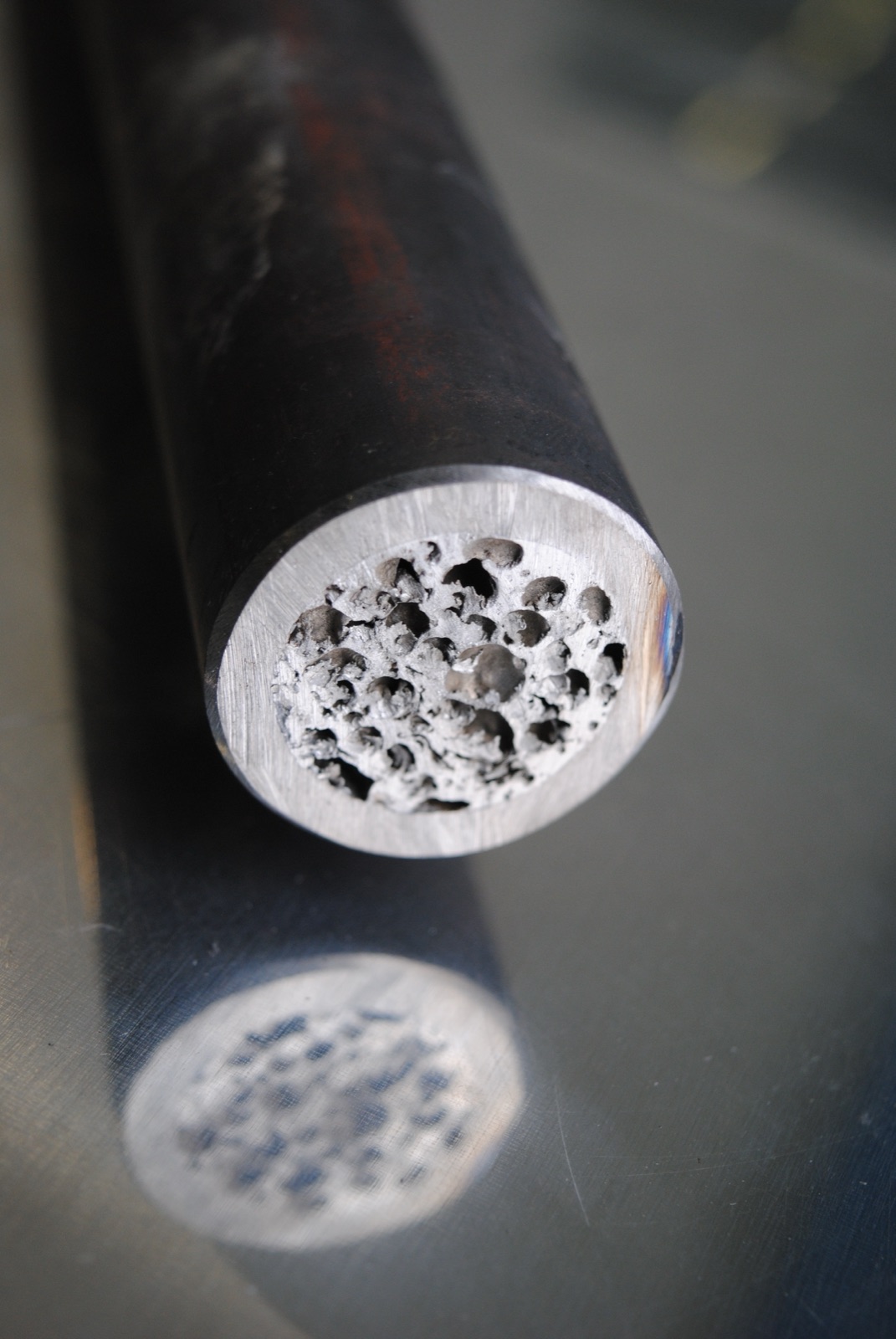 Close-up of the cut end of a dark-coated round profile: in the bright milled end face lies the aluminum foam core with clearly separated pores.