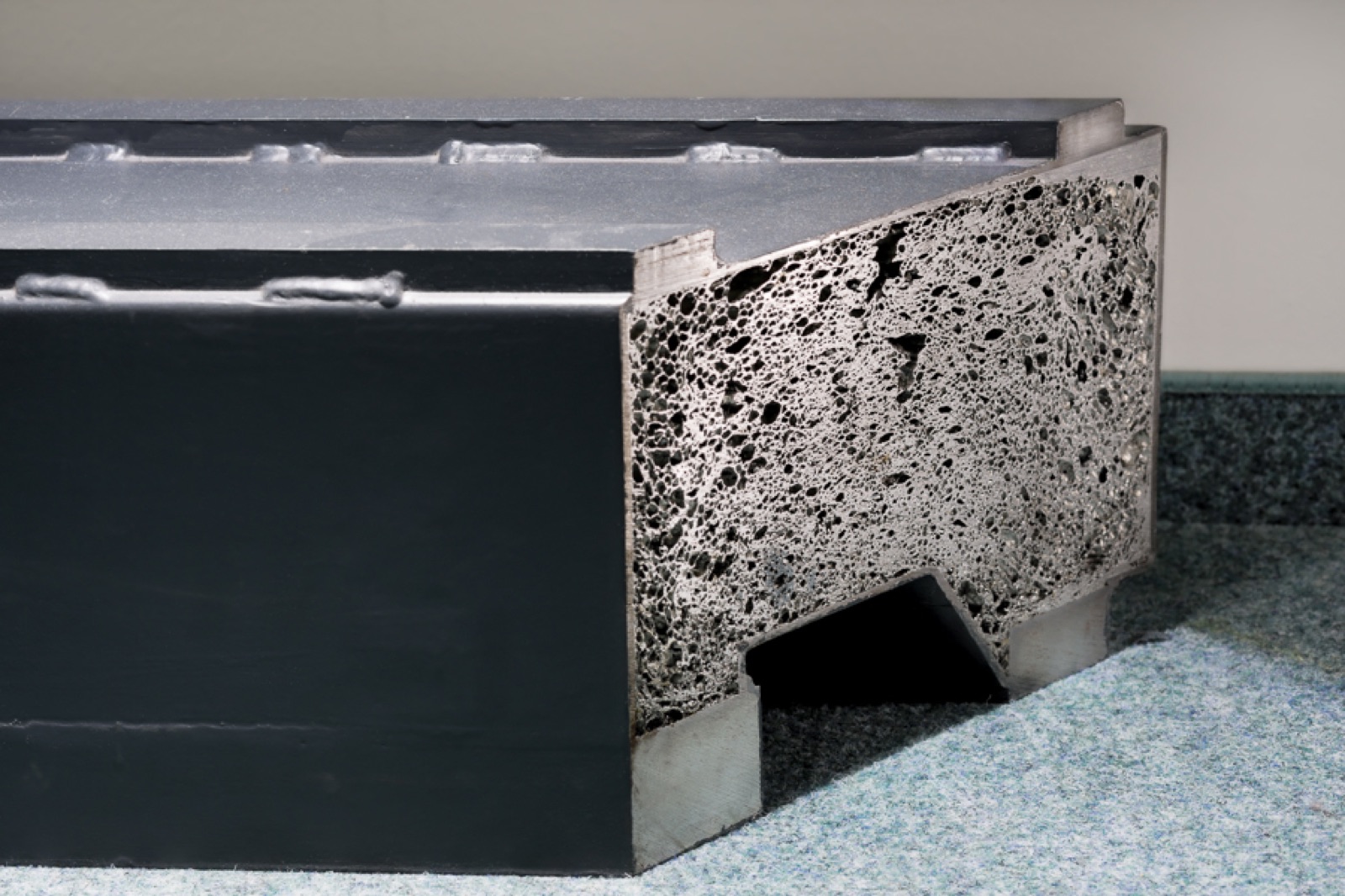 The corner of a large sandwich component with a dark-coated cover layer and welds all round. On the cut end face the aluminum foam core can be seen over the full height of the component.