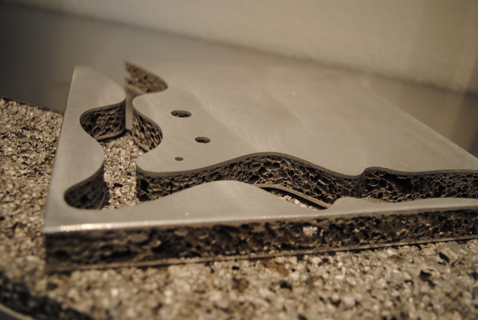 A sandwich panel with a freely cut, wave-shaped contour and several drilled holes; the aluminum foam core is visible at every cut edge. The panel lies on a coarse block of foam.