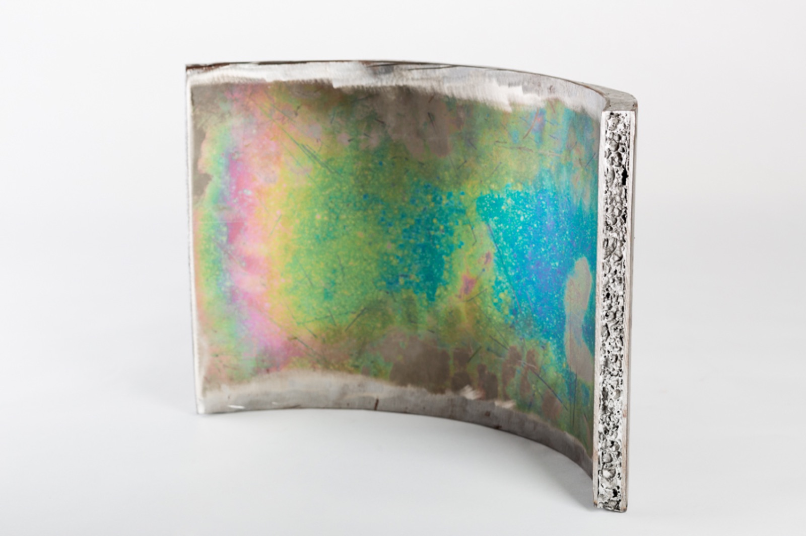 A curved sandwich segment standing on edge; the outer surface shows annealing tints in yellow, green and blue, and at the cut end face the aluminum foam core can be seen between the cover layers.