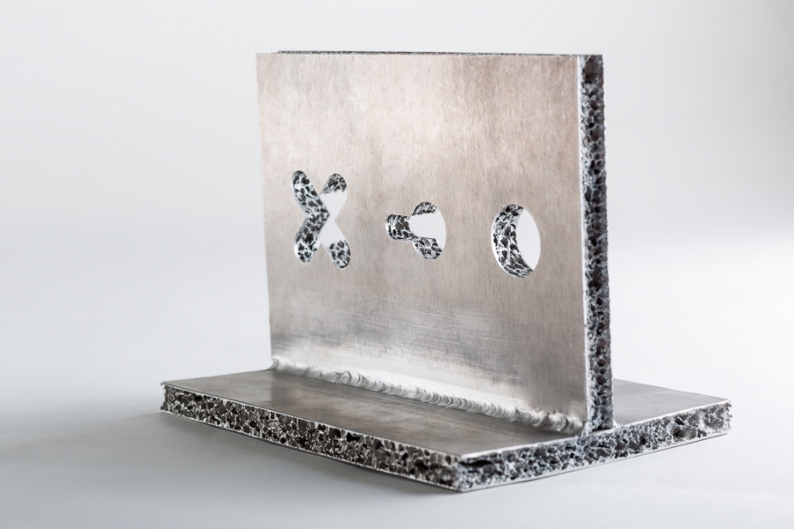 Two sandwich panels welded together at a right angle; the upright panel has three milled cut-outs in the shape of a cross, a keyhole and a circle, and the foam core shows at their cut edges.