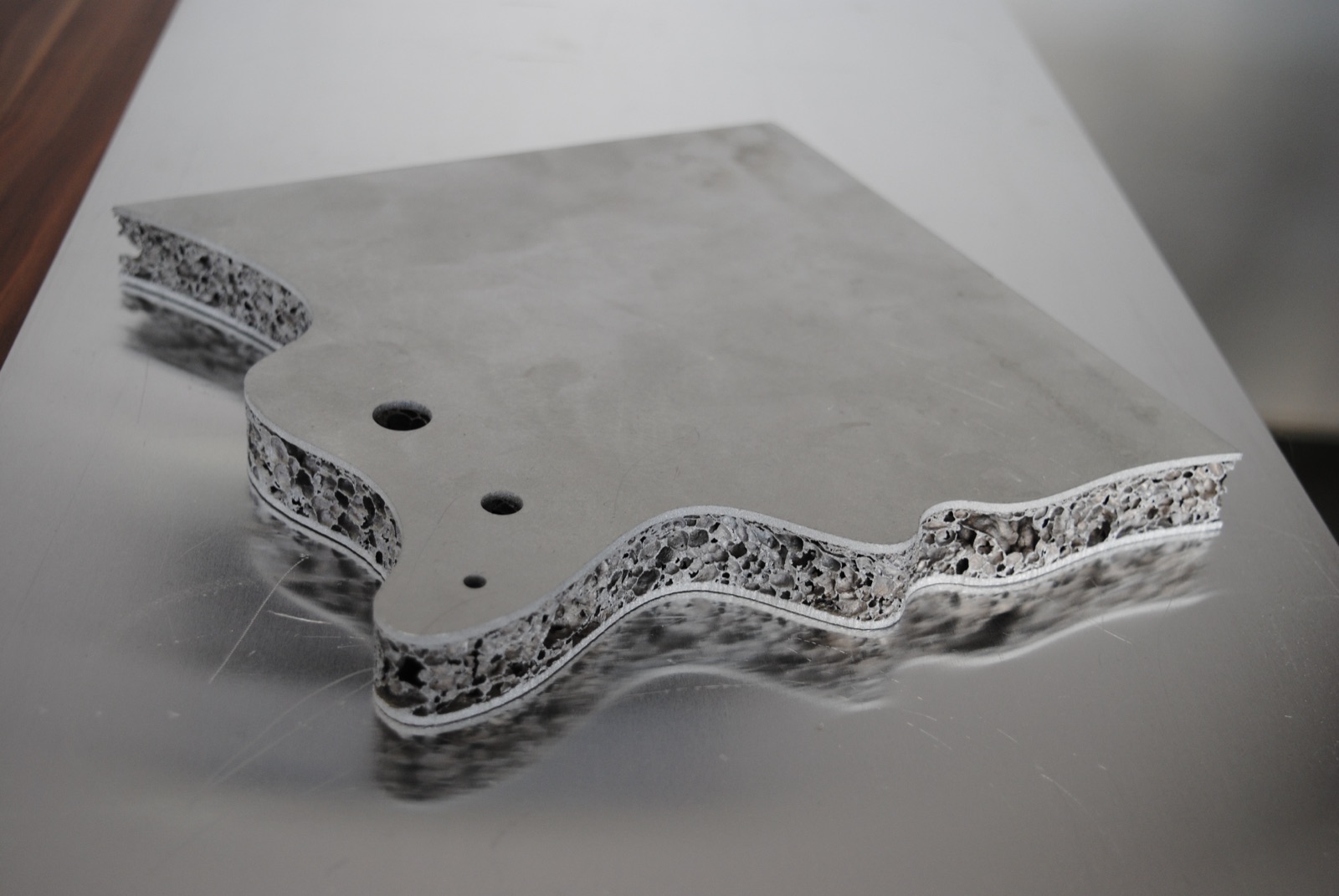 A sandwich panel with a freely cut, wave-shaped outer contour and three holes of different sizes; along the entire cut edge the aluminum foam core lies exposed between the two cover layers.