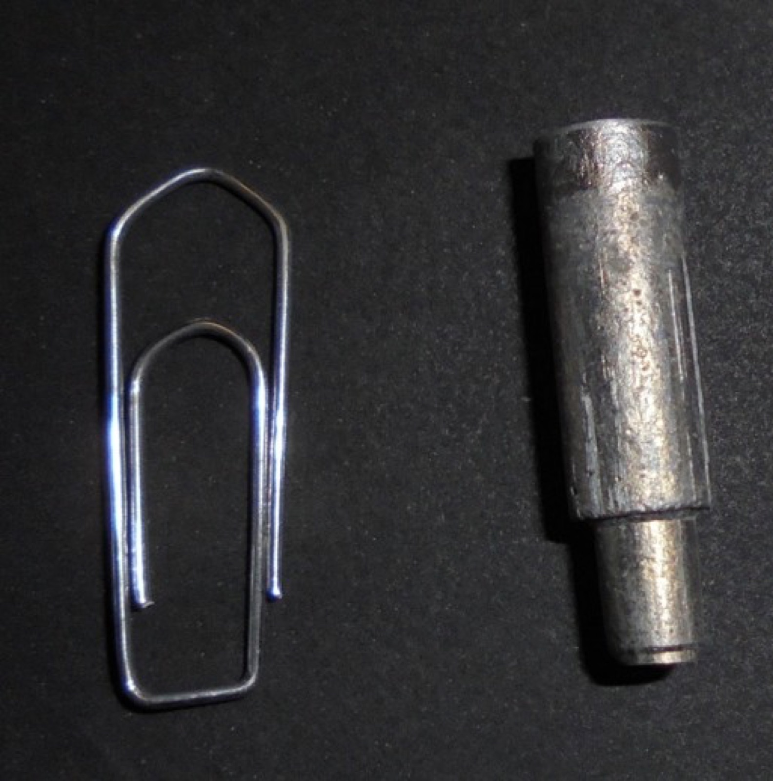 A small cylindrical tappet of aluminum foam with a stepped shank, a paperclip beside it for scale; both lie on a dark surface.
