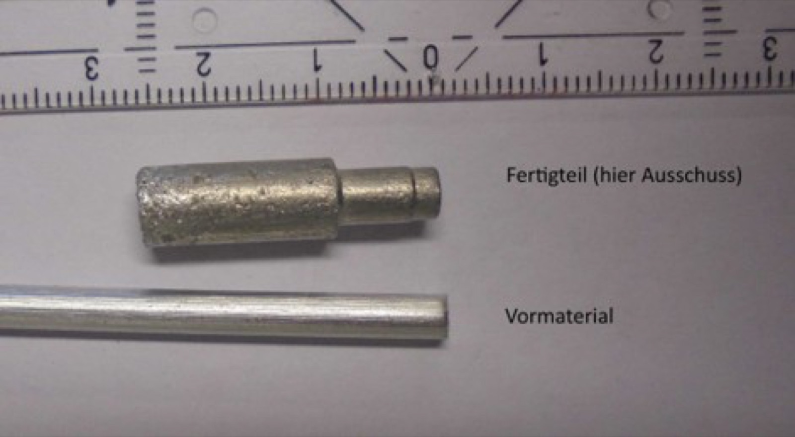 Two parts side by side against a ruler with a centimeter scale: below, a smooth round bar labeled as precursor; above it, a stepped plunger with a foamed surface, labeled as a finished part and marked as a reject.